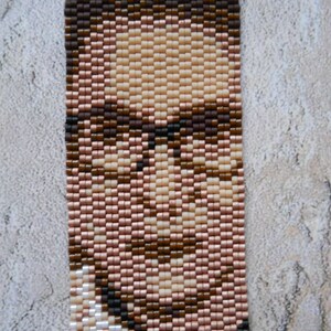 May include: A beaded portrait of a person with brown hair and glasses. The portrait is made with small, colorful beads.