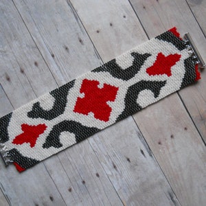 May include: A white, black, and red beaded bracelet with a geometric pattern. The bracelet has silver metal clasps.