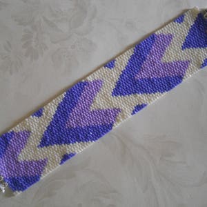 May include: A white and purple beaded bracelet with a geometric pattern. The bracelet has a silver clasp.