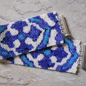 Puede incluir: Un brazalete de cuentas blancas con un patrón geométrico azul y turquesa. El brazalete tiene cierres plateados.