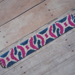 Puede incluir: Una pulsera de cuentas con un patrón geométrico en blanco, azul y rosa. La pulsera tiene forma rectangular con un cierre plateado. El diseño presenta formas entrelazadas.