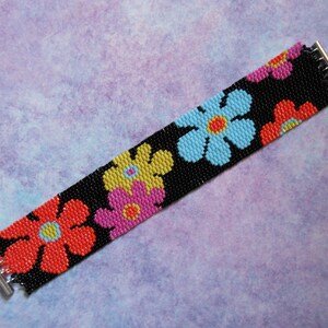 Può includere: Un braccialetto di perline con un motivo floreale. Il braccialetto ha uno sfondo nero con fiori colorati in rosso, giallo, rosa e blu. Il braccialetto è largo circa 4 cm e ha una chiusura argentata.
