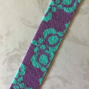 May include: A beaded bracelet with a floral pattern. The bracelet features a repeating design of turquoise flowers on a purple background. The bracelet is rectangular and has silver-colored clasps on each end.