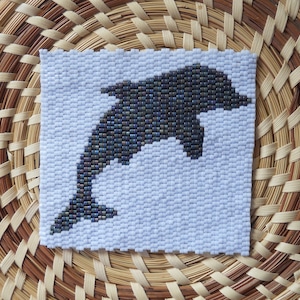 May include: A white beaded square with a black beaded dolphin silhouette. The dolphin is facing right and has a dark blue iridescent sheen.