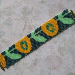 May include: A beaded bracelet with a repeating pattern of yellow flowers with green leaves on a dark blue background. The bracelet has gold metal clasps.