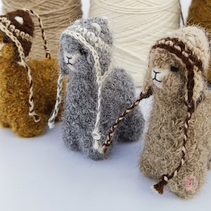 May include: Three felted llama figurines wearing crocheted hats. The llamas are brown, gray, and beige. The hats are brown, white, and beige.