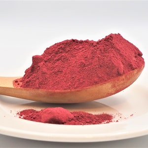 Puede incluir: Una cuchara de madera llena de una buena cantidad de polvo de remolacha roja oscura. El polvo está en un plato blanco con un pequeño montón de polvo derramado en el plato.