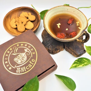 以下が含まれることがあります： 「自然 cafe」と金色で書かれた茶色の箱。「自然 cafe」は「自然カフェ」と訳されます。箱は白い表面に置かれ、緑の葉が周りにあります。白い陶器のボウルは、茶色の取っ手が付いており、茶色の液体と赤いベリーで満たされています。ボウルは木製のスタンドの上に置かれています。