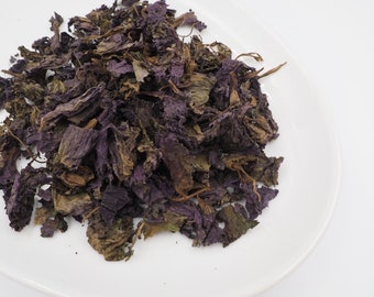 May include: A pile of dried purple shiso leaves on a white plate. The leaves are a deep purple color and have a slightly wrinkled texture.