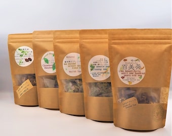 Puede incluir: Cinco bolsas de papel marrón de té mezclado están dispuestas en fila. Cada bolsa tiene una ventana transparente que muestra las hojas de té. Las bolsas tienen etiquetas con texto japonés y descripciones en inglés, incluyendo "Cinnamon L Blended T" y "Pine needle and Ginseng tea."