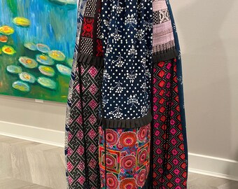 Puede incluir: Una falda larga de patchwork con cintura elástica negra. La falda presenta varias telas estampadas en azul marino, rosa, rojo y blanco. Un detalle de volantes azul oscuro acentúa el dobladillo. La falda se exhibe en un maniquí.