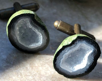 May include: A pair of cufflinks featuring a green and black agate geode with a white interior. The cufflinks are made of metal and have a polished finish.