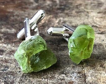 May include: A pair of cufflinks featuring raw, green peridot gemstones set in silver.