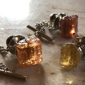 May include: Three tie tacks with different colored gemstones. The gemstones are all cut into different shapes. The tie tacks are made of silver metal.