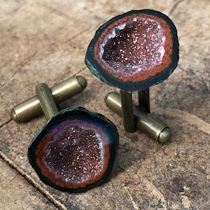 Puede incluir: Un par de gemelos con un diseño de geoda. Los gemelos están hechos de latón y tienen una geoda de color marrón oscuro, verde y rojo con un interior de cristal blanco.