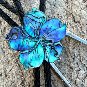 May include: A bolo tie featuring a flower-shaped pendant with iridescent blue and green hues. The pendant is set against a black braided leather cord, with silver-toned tips. The tie is a unique accessory.