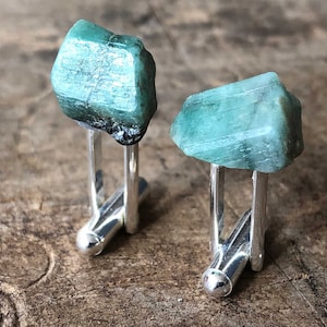 May include: A pair of cufflinks featuring a green emerald gemstone on each. The cufflinks are made of silver and have a polished finish.