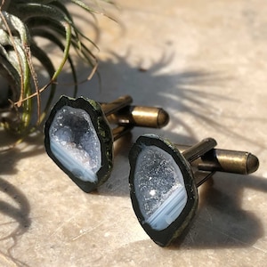 May include: A pair of cufflinks featuring a geode design. The cufflinks are made of brass and have a dark green finish. The geode is a light blue color with white stripes.