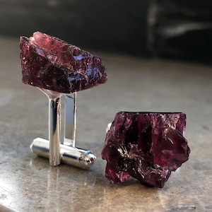 Puede incluir: Un par de gemelos con dos piedras preciosas sin pulir de color morado oscuro engastadas en plata.