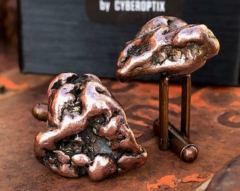 May include: A pair of copper cufflinks with a rough, textured surface. The cufflinks are in the shape of small, irregular rocks. The cufflinks are sitting on a rusty metal surface. The cufflinks are packaged in a black box with the text "WELL DONE goods by CYBEROPTIX".