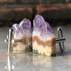 May include: A pair of silver cufflinks featuring natural amethyst crystal formations. The crystals display shades of purple, white, and brown, with a polished silver backing. The cufflinks are set against a blurred brick background.