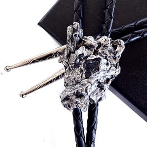 Op de afbeelding: Een zilveren bolo tie met een grote, ruwe, rotsachtige hanger. De stropdas is gemaakt van zwart gevlochten leer.