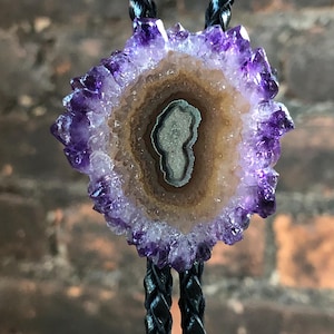 May include: A bolo tie featuring a geode slice with a vibrant purple crystal exterior and a brown and gray interior. The pendant is suspended from a black braided cord, creating a striking accessory.