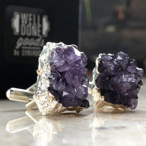 May include: A pair of cufflinks featuring amethyst crystal clusters encased in silver. The amethyst is a deep purple color.