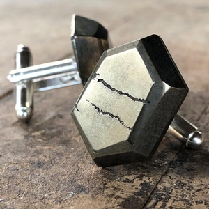 Könnte beinhalten: Ein Paar Manschettenknöpfe mit einer silbernen Metallbasis und einem facettierten Pyrit-Stein. Der Stein ist dunkelgrau mit einem metallischen Glanz und hat einen natürlichen Riss, der durch ihn verläuft.