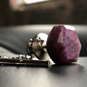 May include: A silver tie tack with a purple gemstone. The gemstone is faceted and has a rough, natural look.
