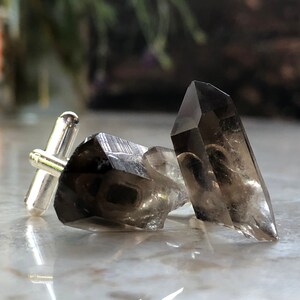 May include: A pair of cufflinks featuring two smoky quartz crystals attached to silver cufflink backs.