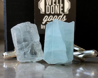 May include: A pair of cufflinks featuring two light blue aquamarine crystals set in silver.