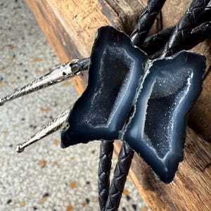 May include: A bolo tie featuring two dark blue geode slices with a crystalline interior. The geode stones are attached to a braided black leather cord. The silver-colored tips have an ornate design.
