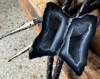 May include: A bolo tie with two dark blue geode slices and a crystalline interior. The geode stones are attached to a braided black leather cord. The silver tips have an ornate design.