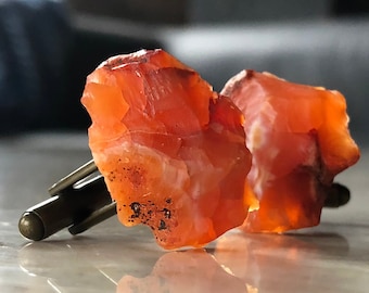 May include: A pair of cufflinks made from natural carnelian stone. The cufflinks are a deep orange color with a rough, natural texture. The cufflinks are attached to a silver metal base.