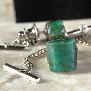 May include: Three emerald green gemstone tie tacks with silver metal settings and chains. The gemstones are rough cut and have a natural, unpolished look.