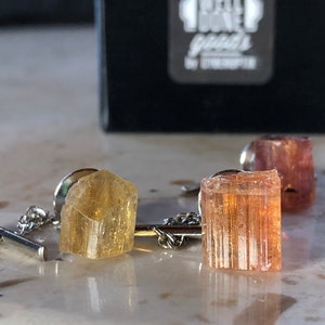May include: Two gold and orange crystal tie tacks with silver chains. The tie tacks are set against a white background.