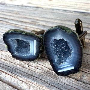 May include: A pair of cufflinks featuring irregular, sliced agate stones with dark, crystalline centers and banded gray edges. The stones are mounted on dark metallic hardware with a toggle-style backing, showcasing natural mineral textures.