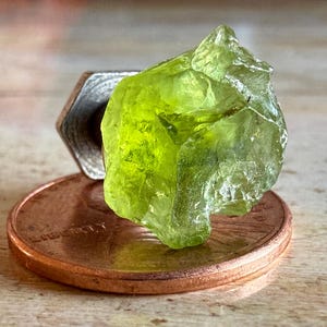 May include: A rough, lime-green peridot gemstone rests on a copper penny. The gemstone is irregularly shaped with a crystalline structure. A silver-colored, hexagonal object is partially visible behind the gemstone.