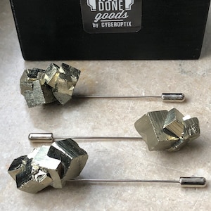 Könnte beinhalten: Drei goldfarbene Pyrit-Kristall-Reversnadeln mit silberfarbenen Metallnadeln. Die Nadeln sind in einer Reihe auf einer grauen Oberfläche angeordnet. Der Text "WELL DONE goods BY CYBEROPTIX" ist auf einer schwarzen Box im Hintergrund sichtbar.