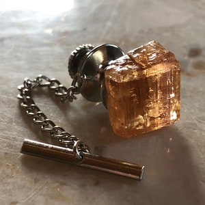May include: A gold-toned tie tack with a clear, faceted, orange crystal. The crystal is attached to a silver chain and a silver bar.
