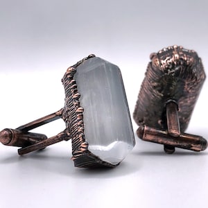 May include: A pair of cufflinks with a white selenite crystal set in a copper frame. The cufflinks have a textured design and are a unique and stylish accessory.