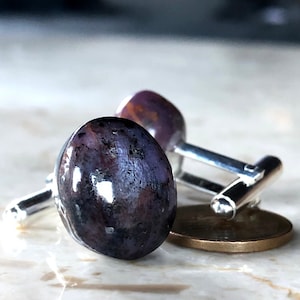 May include: A pair of silver cufflinks featuring a dark purple and silver gemstone. The cufflinks are oval shaped and have a polished finish.