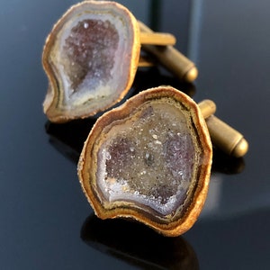 May include: A pair of cufflinks featuring a geode design. The cufflinks are made of brass and have a natural, brown finish. The geode is a light purple and white color.