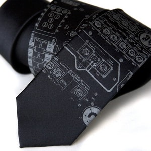 Può includere: Una cravatta nera con una stampa di circuito stampato di colore grigio argento. La cravatta presenta varie etichette, tra cui "POST LANDING", "VENT HIGH", "COAS POWER" e "WASTE MAN".