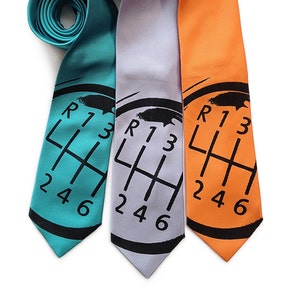 Könnte beinhalten: Drei farbige Krawatten in Türkis, Grau und Orange. Jede Krawatte zeigt ein schwarzes Muster eines Autoschalthebels mit den Zahlen R, 1, 3, 2, 4 und 6. Die Krawatten sind oben aufgerollt.