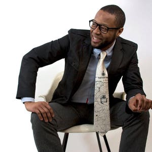 May include: A man wearing a black suit jacket and a white shirt with a patterned tie. The tie has a map design with a compass and a ship.