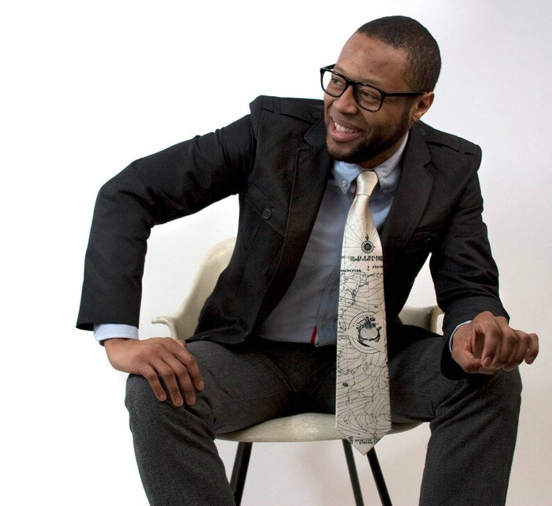May include: A man wearing a black suit jacket and a white shirt with a patterned tie. The tie has a map design with a compass and a ship.
