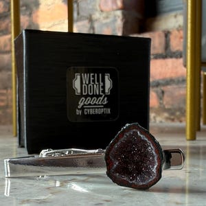 May include: A silver-toned metal tie bar featuring a dark, textured, crystalline geode stone accent. Behind the tie bar sits a black square box with the white printed text WELL DONE goods by CYBEROPTIX centered on the front face of the box.