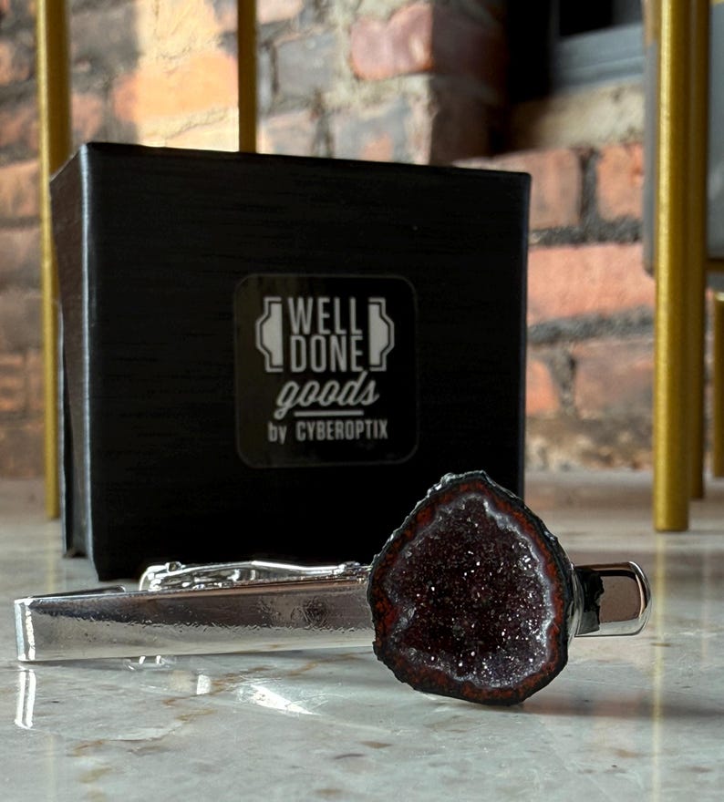 May include: A silver-toned metal tie bar featuring a dark, textured, crystalline geode stone accent. Behind the tie bar sits a black square box with the white printed text WELL DONE goods by CYBEROPTIX centered on the front face of the box.